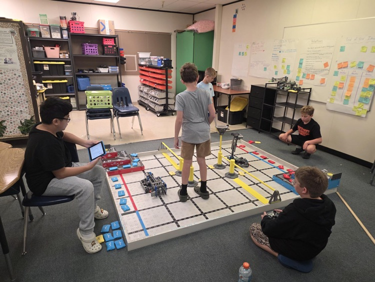 students sitting around robotics field and one standing on the field 