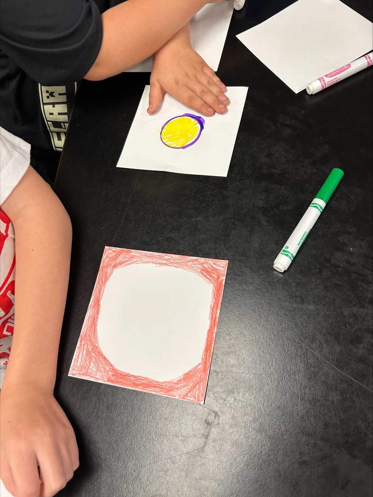 Two people sit at a table with colored markers and paper. One draws a circle on paper with yellow marker.