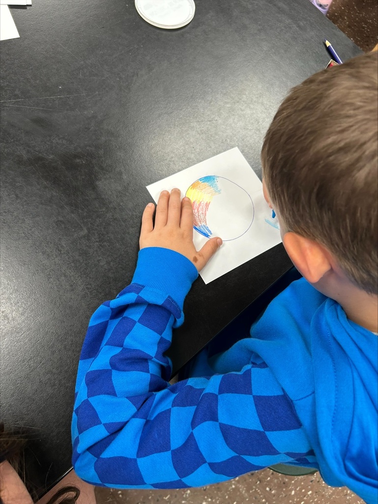 A boy in a blue checkered long-sleeve shirt is drawing a circle on a piece of paper.