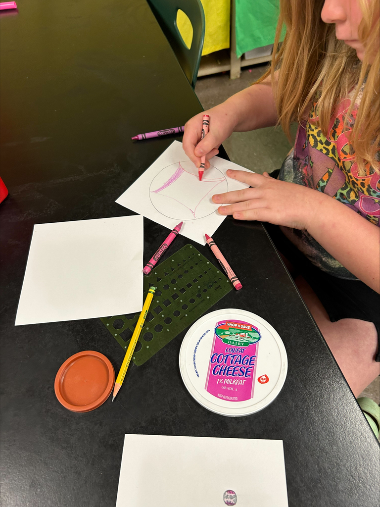 A child seated at a table draws on paper with colored markers, surrounded by paper, a plate, and a pencil.