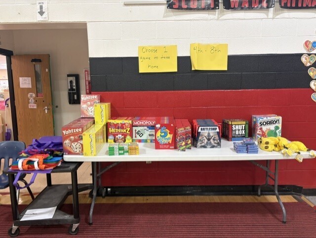 Table with various items, including toys, books, and stuffed animals, on a white tablecloth. Red and white walls, yellow papers, and a door in background.