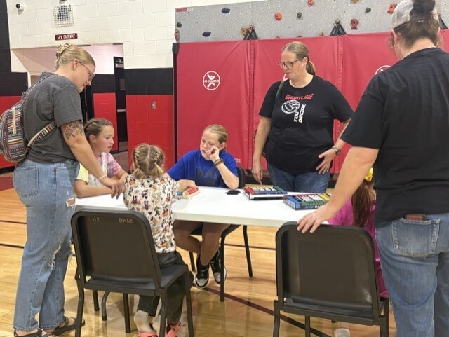 A group of people, including children, in a gymnasium with a red wall. One person sits at a table.