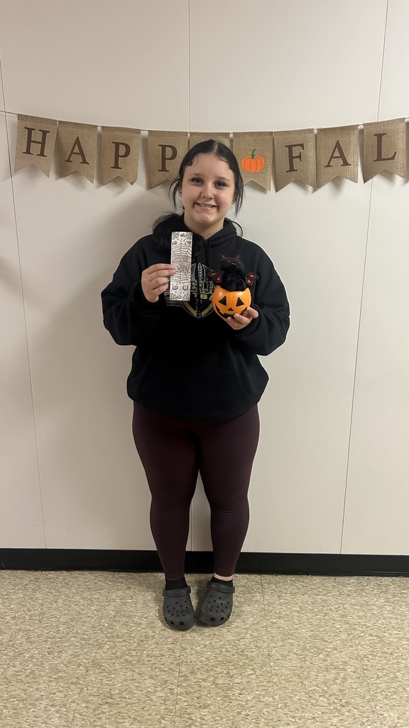 A person stands smiling with a small orange pumpkin and a card in hand in front of a white wall.