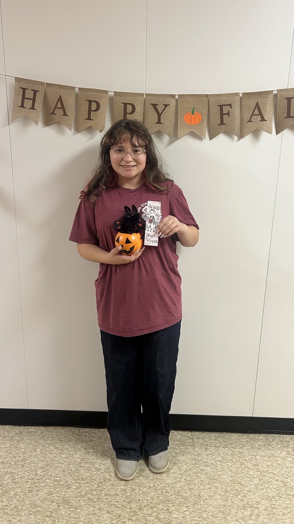 A student stands with a pumpkin and a white object in hand against a white wall with a banner.