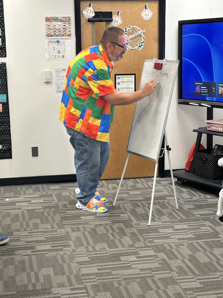 A person stands at a whiteboard writing with a marker. A television is behind him.