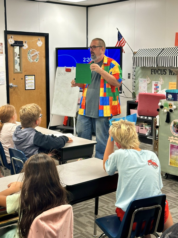 Man in colorful shirt stands at front of classroom with large green board. Four children sit at desks facing him.