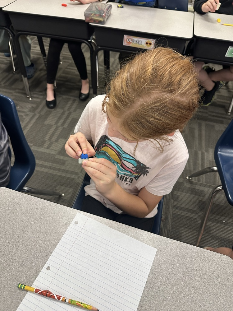 A young student wearing a white shirt sits at a desk, using a tool to sharpen a pencil.
