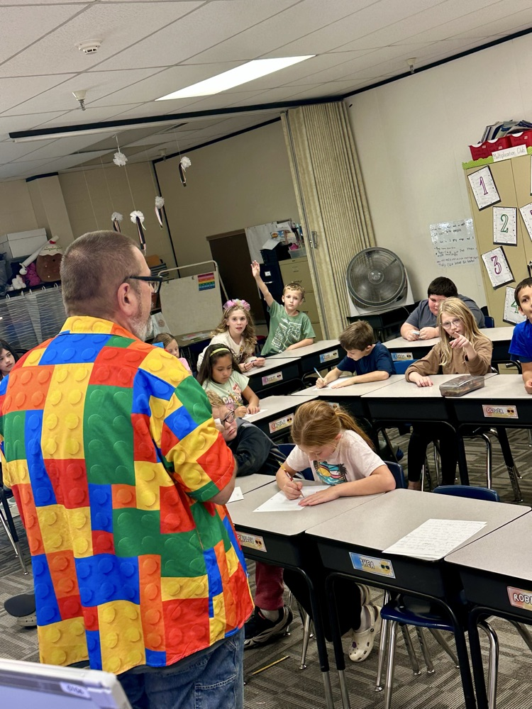 Teacher standing in front of a classroom of children seated at desks writing, with some raising their hands.