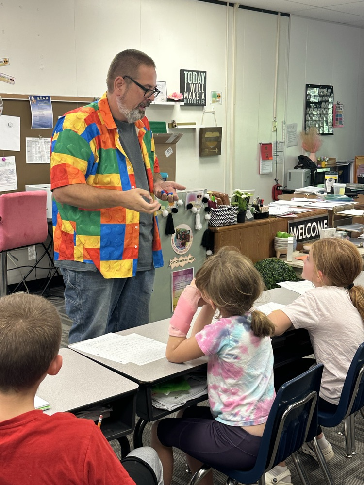 A man in a colorful outfit stands before a classroom table with three students sitting at it.