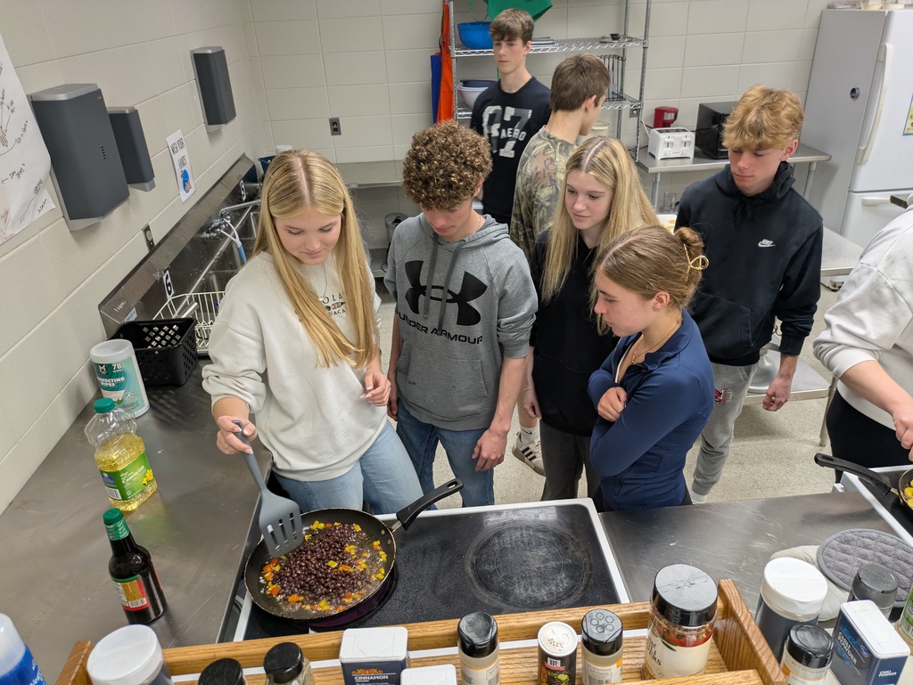 Students gather around as one student makes the dish in a pan on the stove 
