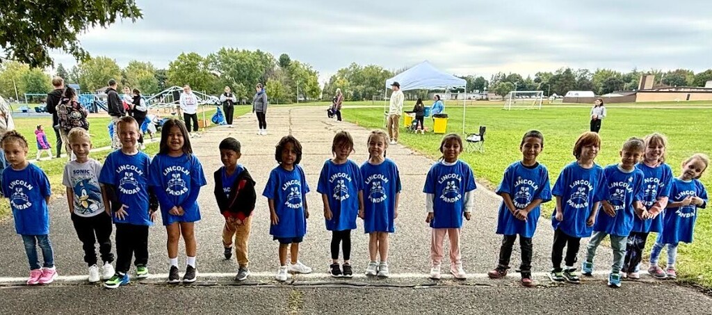 A line of children in matching blue shirts and sneakers stands on a road with green grass.