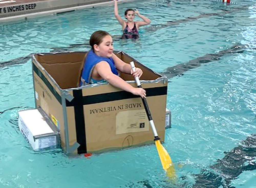 A student in swim suit and life vest confidently paddles a cardboard boat she made out of boxes and waterproof duck tape across the surface of the Whitney Point swimming pool.