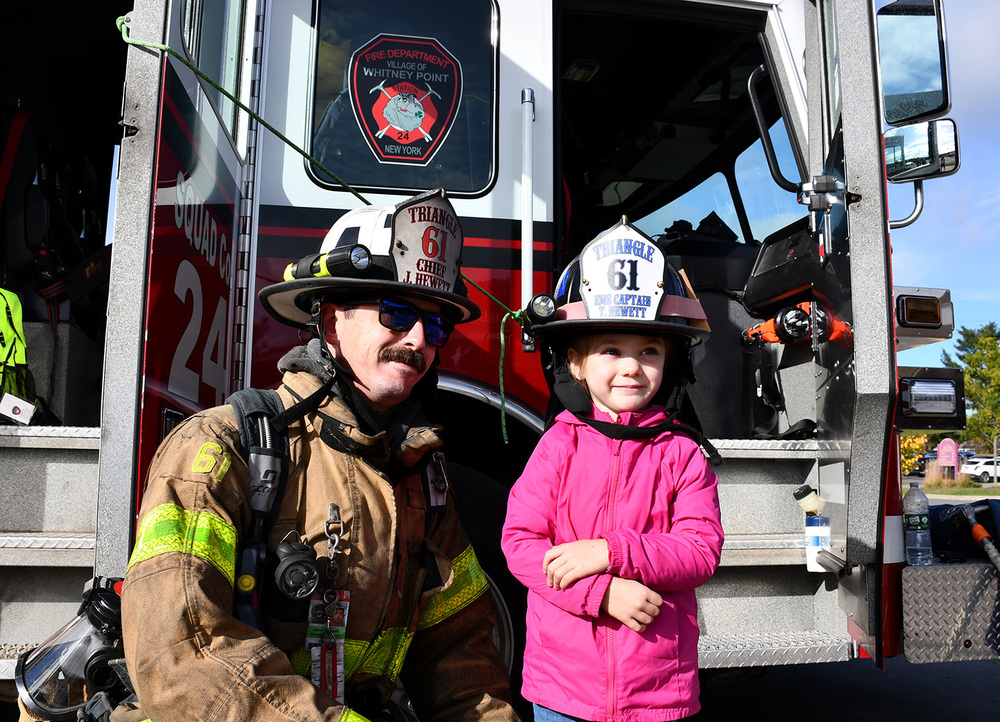 A fire fighter wearing gear and a helmet poses next to Pre Kindergarten student also wearing a fire helmet.