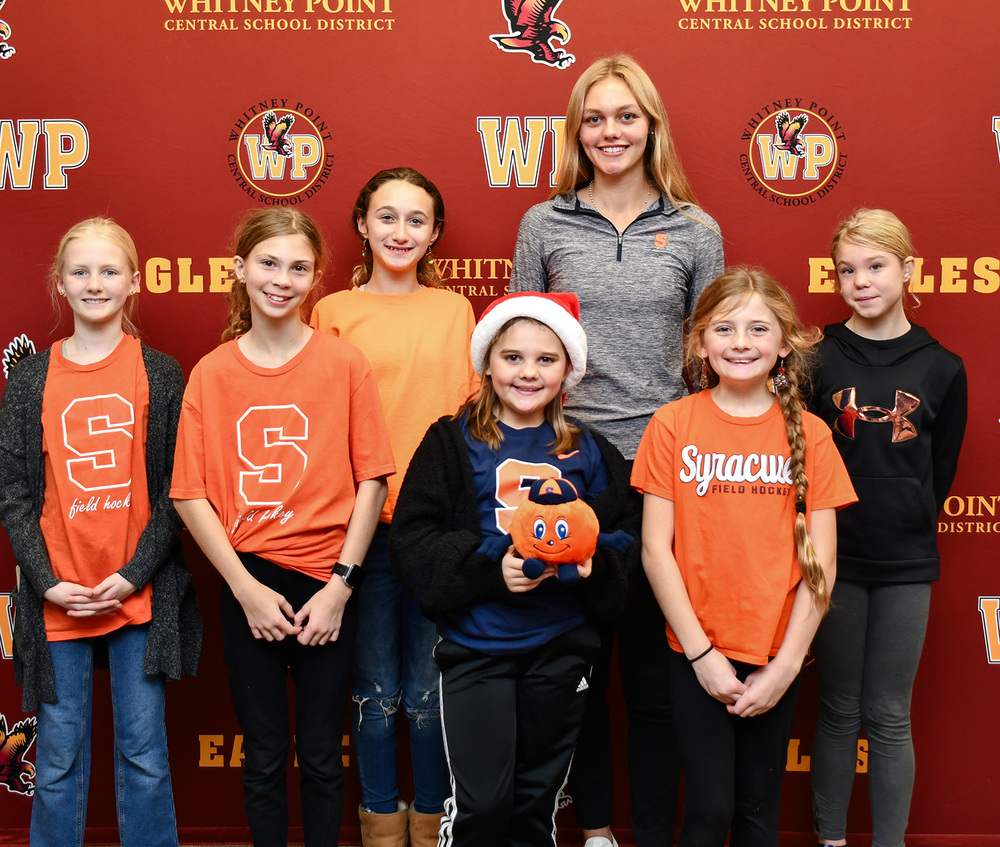 Whitney Point High School senior Genevieve Huston stands in front of the Eagle wall after her Letter of Intent signing with a group of younger students she coaches in field hockey.