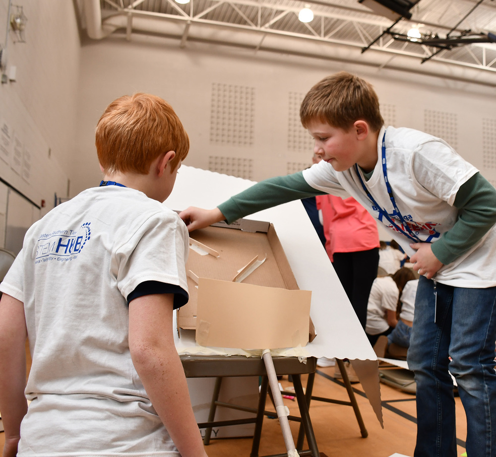 A Tioughnioga Riverside Academy student stands on a chair to position a chute on the Rube Goldeberg machine he is constructing with teammates as another student watches.