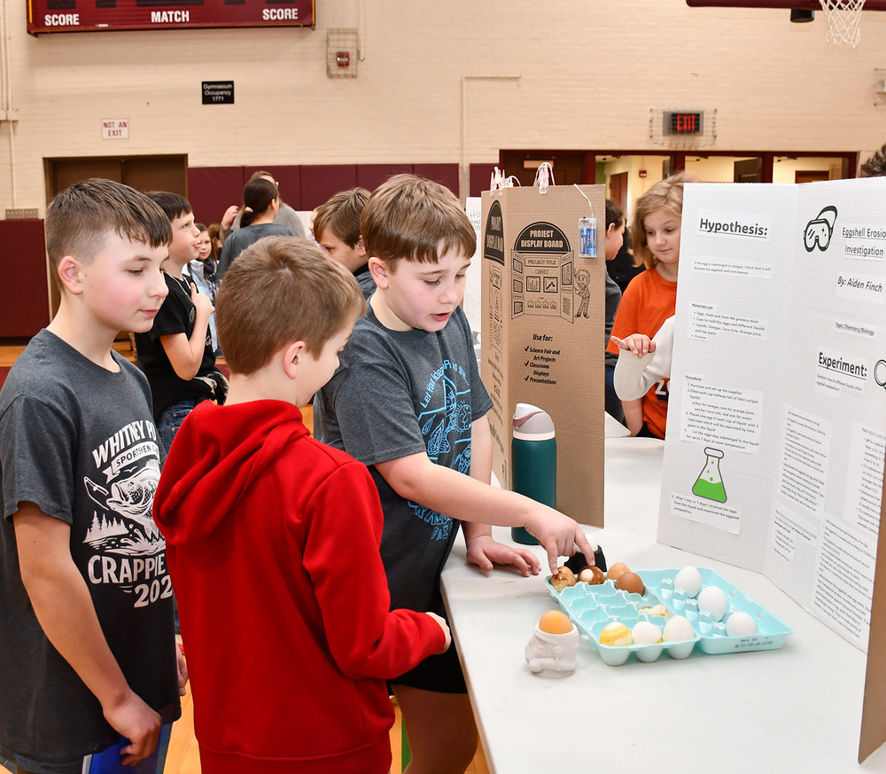 A Tioughnioga Riverside Academy student points to an open carton of eggs as he explains his science fair entry "Eggshell Erosion Investigation" to two Caryl Adams students.