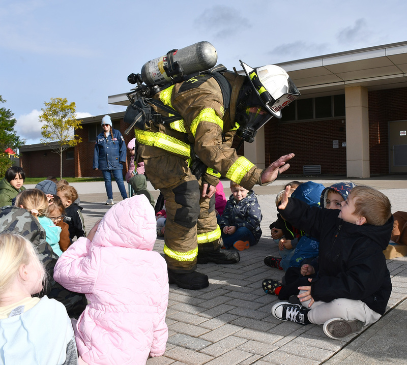 A fire fighter in full gear high fives a young child sitting in front of his school during a fire safety lesson.