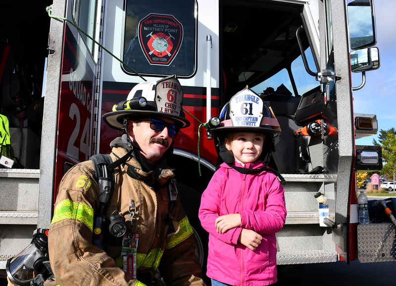 A fire fighter wearing gear and a helmet poses next to Pre Kindergarten student also wearing a fire helmet.