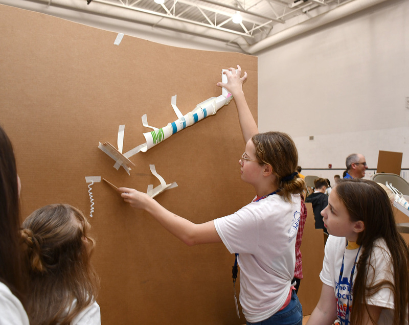 A T R A student holds up a tube that her team is taping to a large section of cardboard to construct their Rube Goldberg machine.