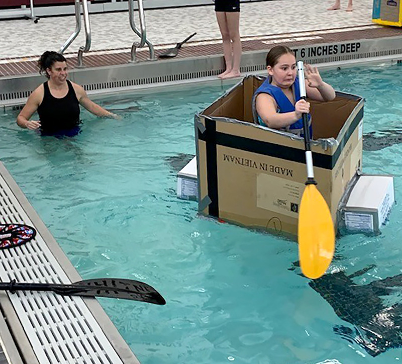 A student with a startled expression on her face begins to paddle her cardboard boat over the swimming pool's surface.