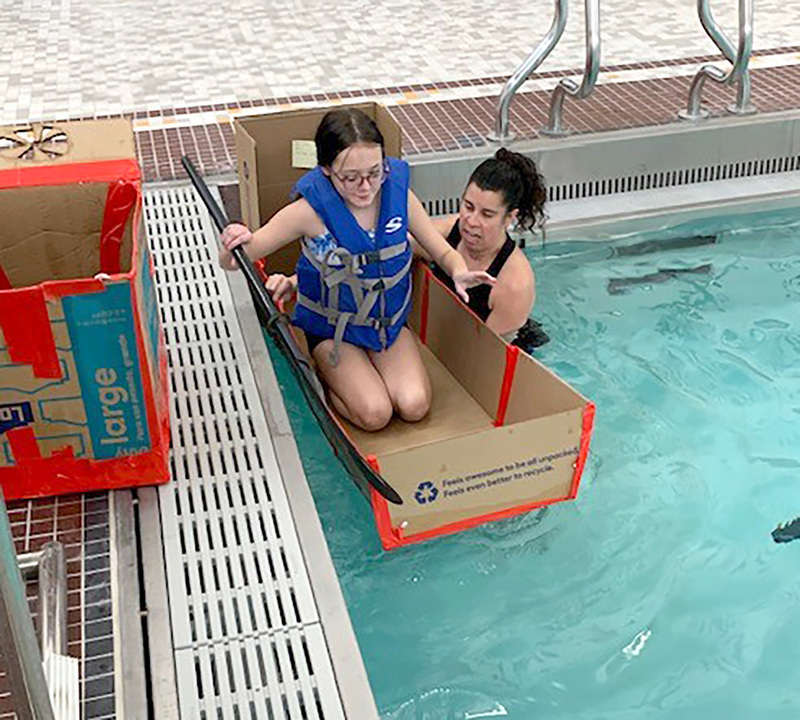 A teacher in the pool steadies another cardboard boat as a student gingerly gets in holding her paddle.