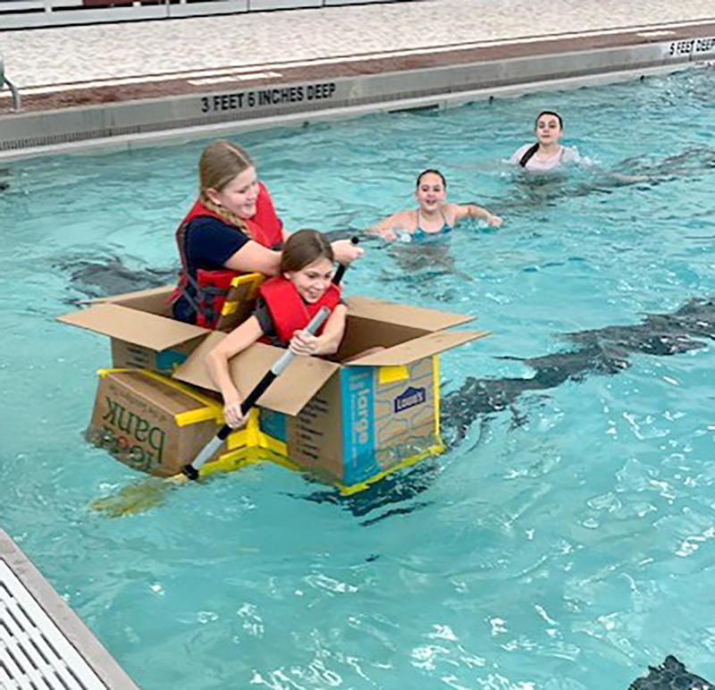 Two students paddle their cardboard vessel across the surface of the pool.