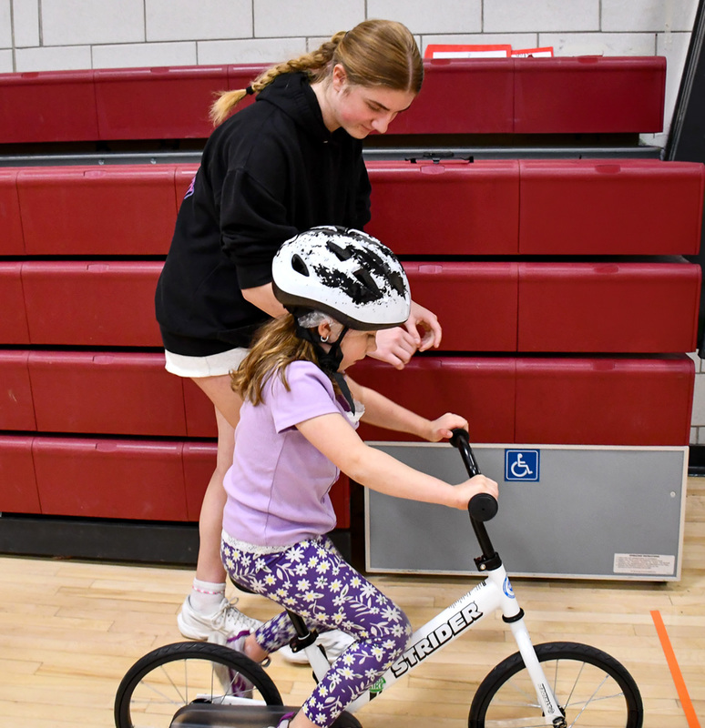 A Whitney Point High School ninth grader walks alongside a first grader as she rides a bicycle during a lesson in her physical education class.