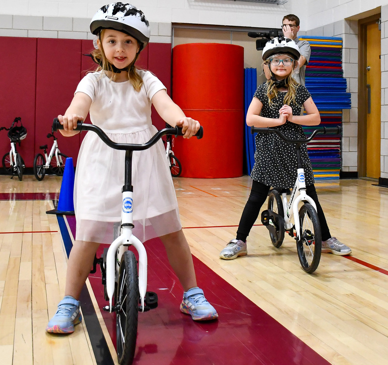 Two Whitney Point first graders pose on their bicycles in physical education class in the gym at Caryl Adams Primary.