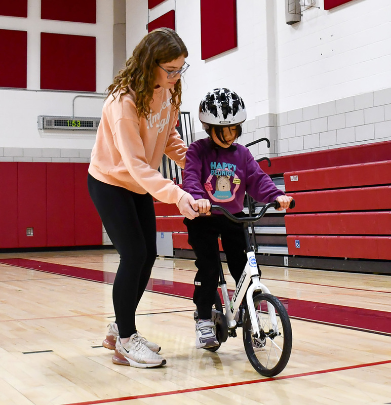 A Whitney Point High School ninth grader steadies a first grader learning to ride a bike in gym glass with one gentle hand at her back and the other helping steady the bicycle's handlebars.
