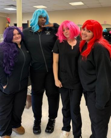 Four Cosmetology students wearing brightly colored wigs smiling in the WT salon.