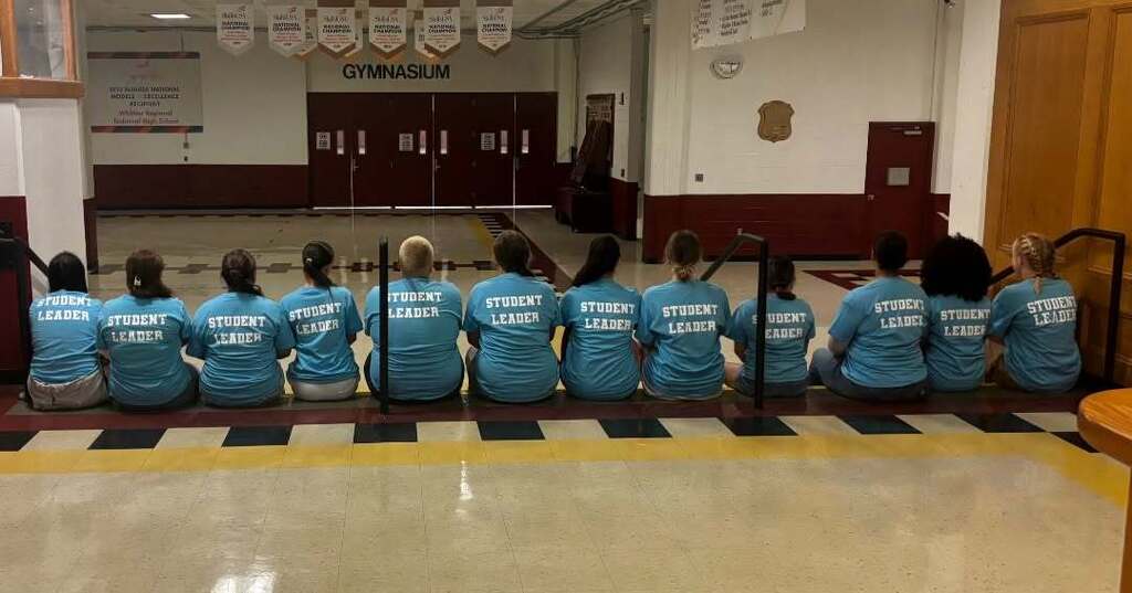 Student Leaders sit on the top step in the main lobby with their backs to the camera so we can see "Student Leader" on their blue shirts.