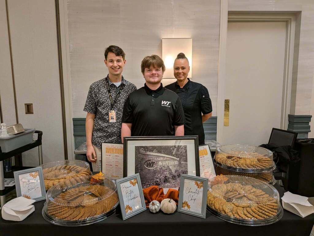 2 staff and 1 student stand behind a table of cookies