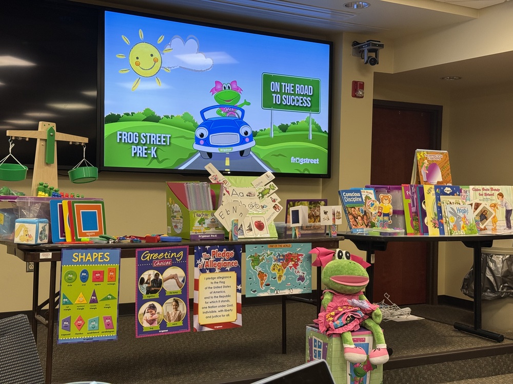 A Frog Street Pre-K curriculum display featuring colorful educational materials, children’s books, posters, classroom resources, and a large frog mascot. A presentation screen in the background shows a cartoon frog driving a car with the words “Frog Street Pre-K” and “On the Road to Success.”