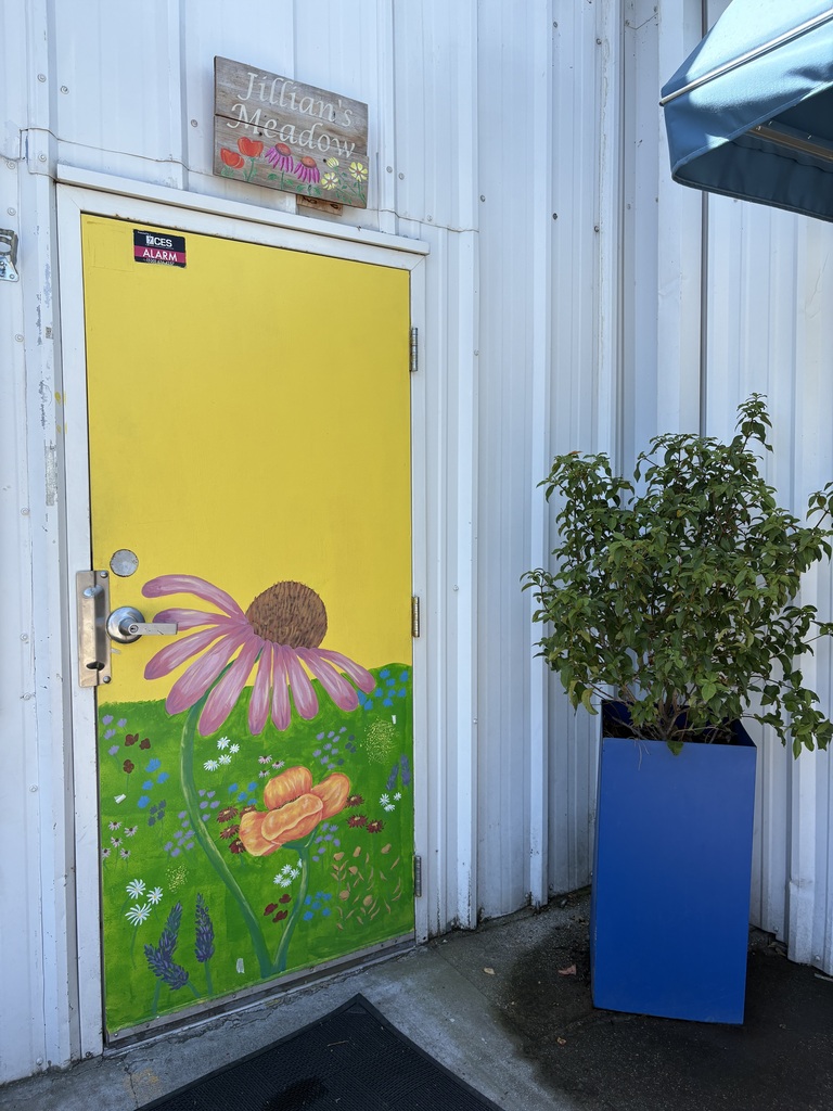 Here is a photo of a yellow door with a bunch of different flowers painted on it. This room is called Jillian's meadow to honor one of our founders. This room serves as our meeting room.