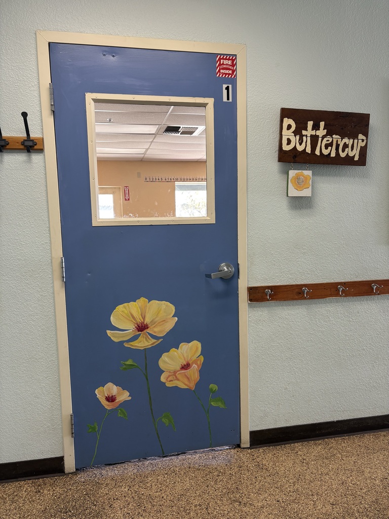 A photo of our Buttercup classroom door for one of our Kinder and 1st grade classrooms. The photo shows a blue door with buttercups painted on it with a sign that says Buttercup on the wall.