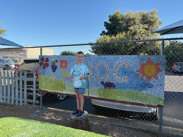 One of our younger students standing in front of a glass mosaic that was made at our Music Festival.