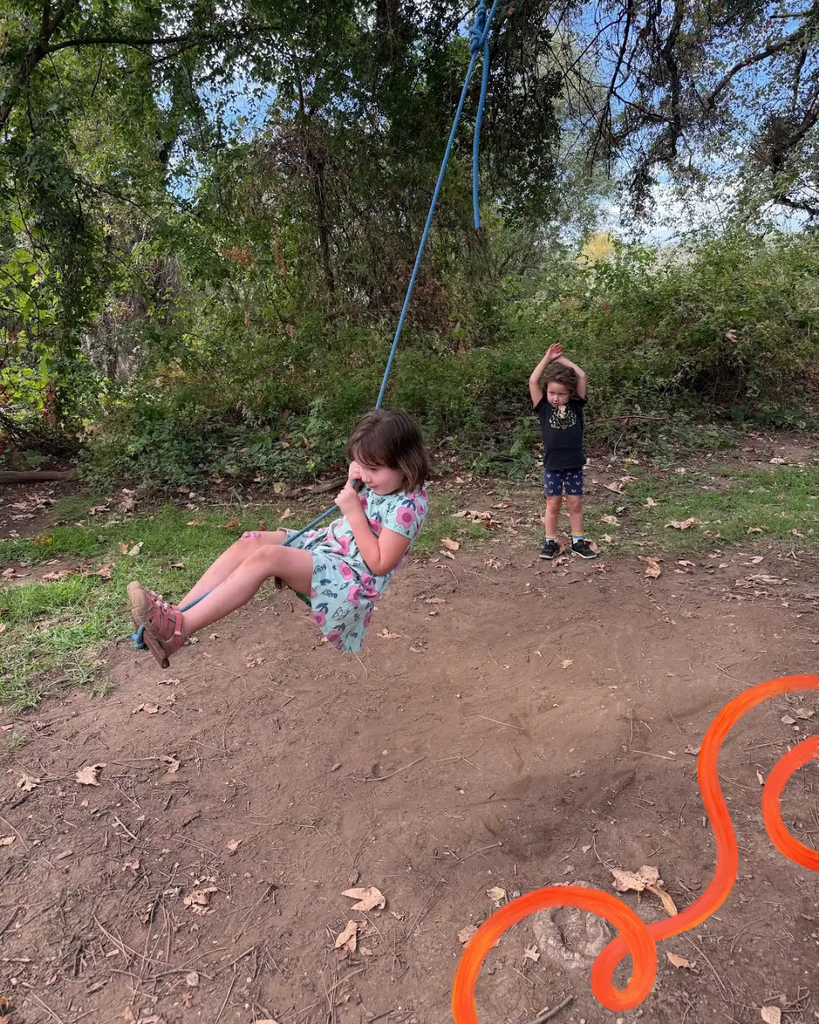 Here is a photo of two TK students swinging on a rope swing that they found. In the phot one student is swinging from the swing attached to a tree and the other student is in the background cheering her on.