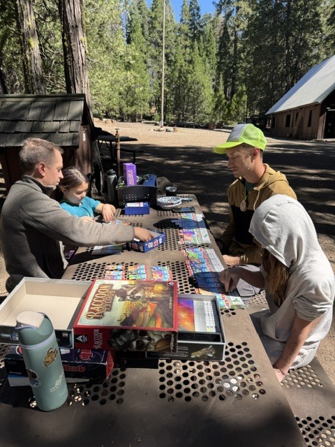 A photo of two students and two parents sitting at a picnic table. They are all playing a card game together under the trees.