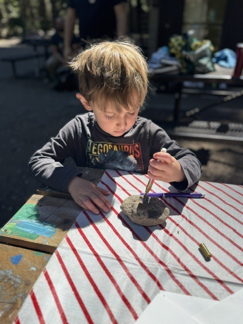Here is a photo of a student sitting at a picnic table. He is painting a rock.