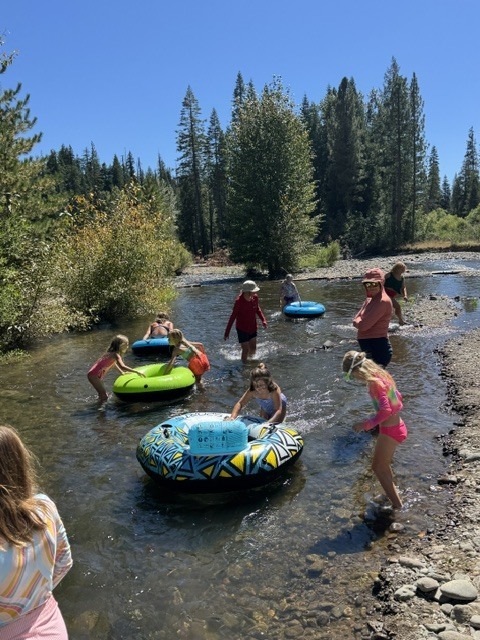 Here is a photo of a bunch of students playing in the creek with inflatable tubes. It is a clear, sunny day and there are lots of trees in the background around the edge of the creek.