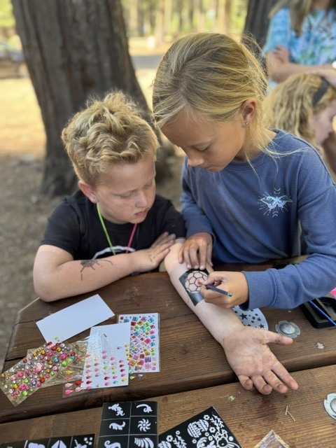 A photo of one student drawing with a stencil on the forearm of another student. They are both sitting at a picnic table and are surrounded by stickers and art supplies.