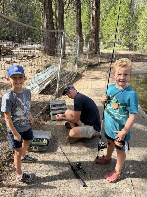 Two children are holding fishing poles with a parent volunteer who is getting lures ready from a tackle box. All three of them are beside either a pond or the edge of a creek.