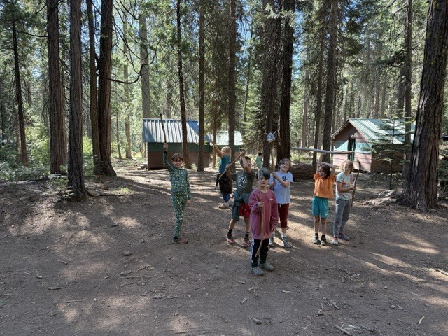 A photo of a group of students playing with big sticks in front of some cabins. The cabins and the students are both surrounded by a bunch of trees.
