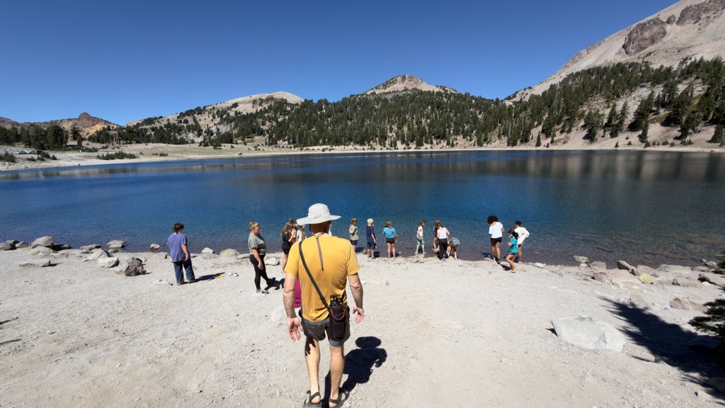 Here is another photo of students on the hike to the alpine lake. There is a larger group of students hanging out on the bank of the lake. Similar to the last photo, there are mountains, trees, and blue sky in the background.