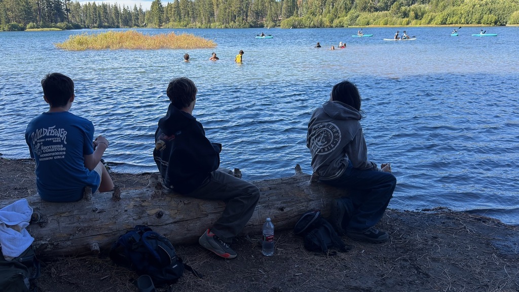 This photo shows 3 students sitting on a log on the shore of the lake in the foreground. In the background, is a bunch of students swimming, kayaking, and paddleboarding.