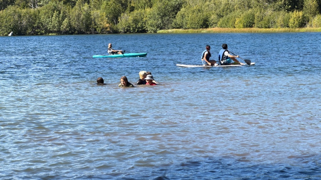 Here is a photo of students swimming, kayaking, and paddleboarding in Manzanita Lake. There are a group of 4 students swimming, 2 students on a paddleboard, and 1 student kayaking in the back.