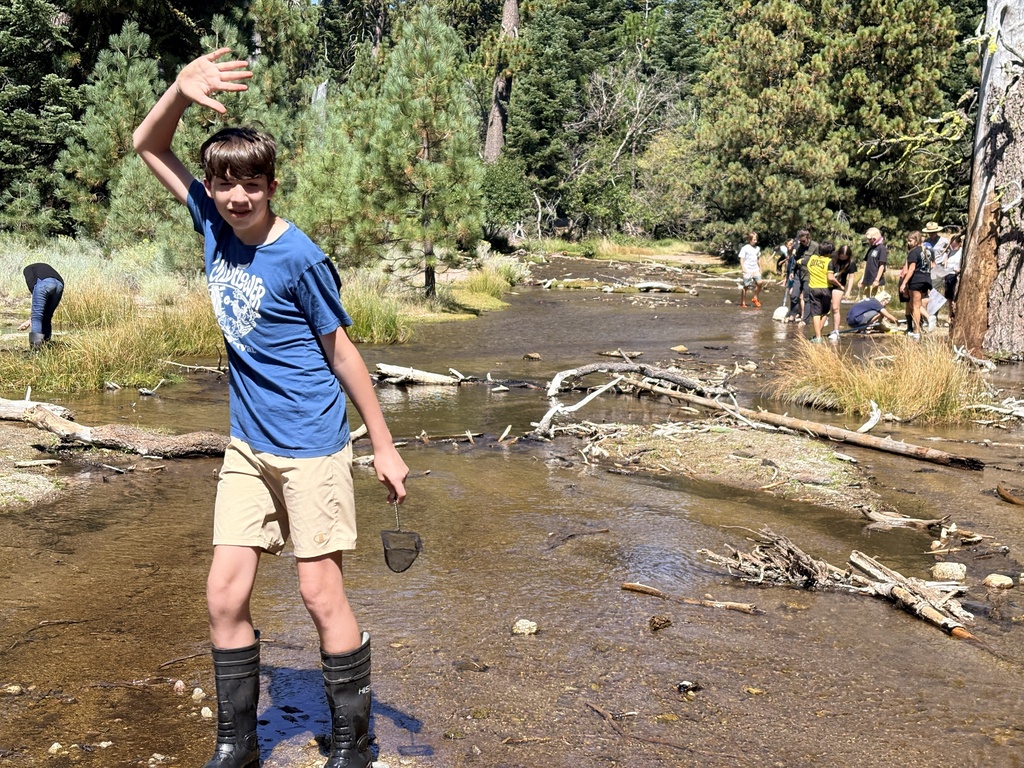This photo shows a students standing in the foreground waving at the camera. He is standing in a creek and exploring the wildlife. In the background is a larger group of students who are also standing in the creek and learning about the wildlife.