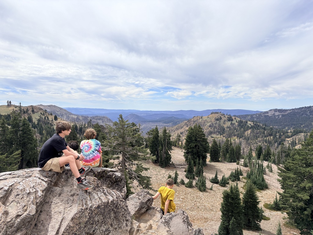 Here is a photo of three students sitting on top of a rock looking out at a view of mountains and trees.