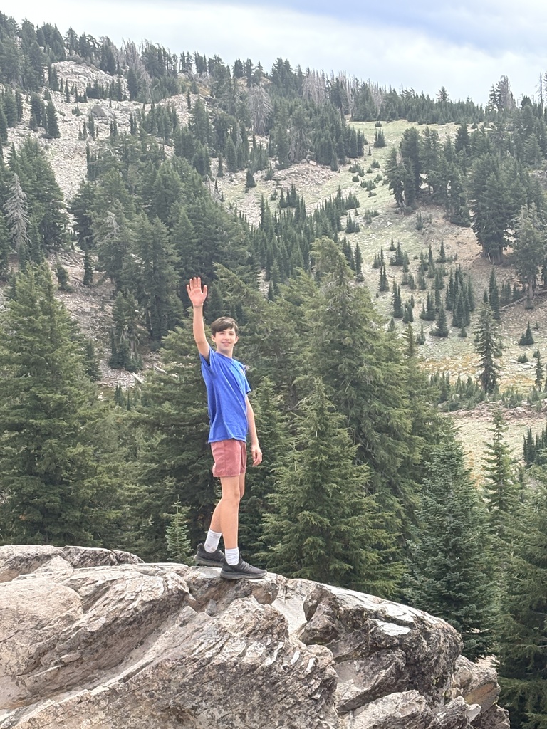 Here is a photo of a student standing on top of a rock outcropping, waving at the camera. In the background is a hill with lots of trees. 