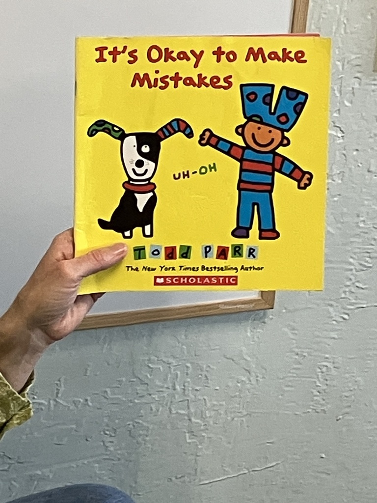 Here is a photo of the book titled "It's Okay to Make Mistakes." The entire cover of the book is yellow and in the middle is a drawing of a dog and a boy. The dog (on the left side) has two socks on his ears and the boy (on the right side) has a pair of pants on his head. The author name and and publishing house are towards the bottom.